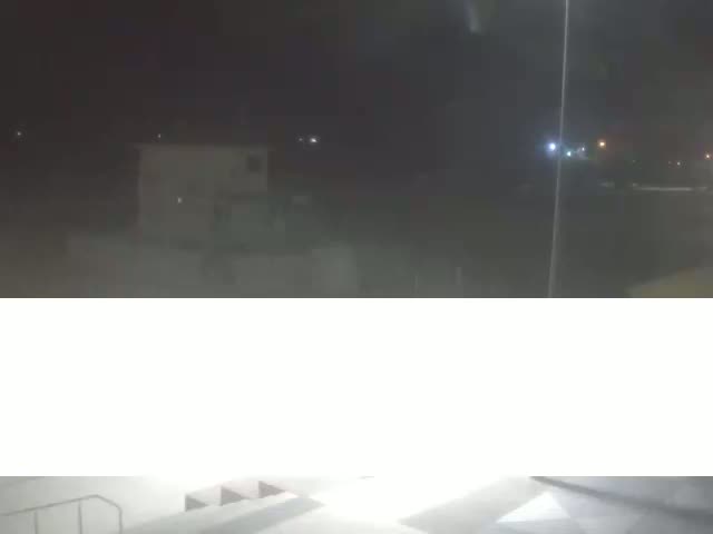 A nighttime, hazy view shows a building and distant lights, with a brightly lit concrete area in the bottom.