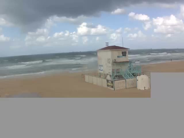 A white lifeguard station with light blue stairs stands on a sandy beach beside a choppy ocean under a partly cloudy sky with both dark and light clouds.