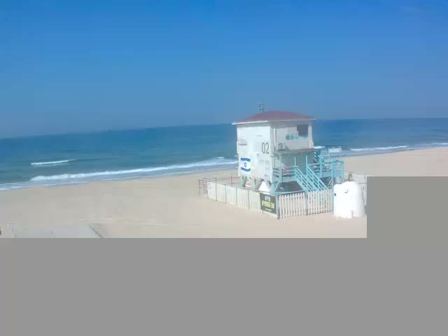 A white lifeguard station with light blue stairs stands on a sandy beach beside a choppy ocean under a partly cloudy sky with both dark and light clouds.