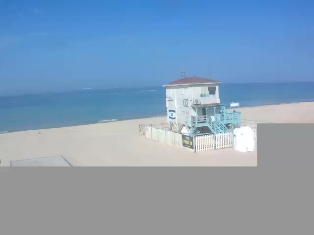 A white and teal lifeguard tower stands on a sandy beach beside a calm blue ocean under a clear blue sky.