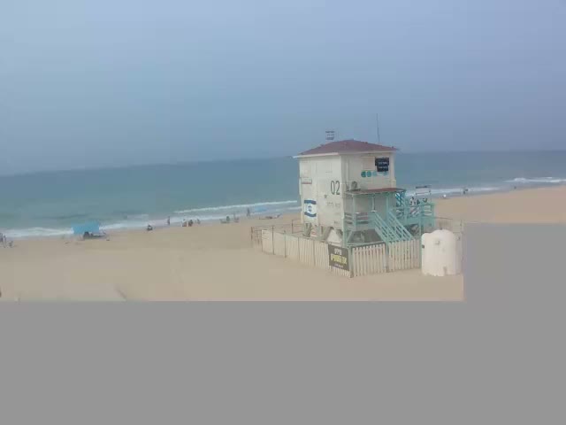 On an overcast day, a multi-story white lifeguard tower with blue stairs stands on a wide sandy beach where people are scattered along the shore and in the calm, blue-green ocean.