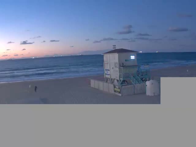 A sandy beach features a white and light blue lifeguard tower, gentle ocean waves, and several distant ships on the horizon, all under a clear, twilight sky transitioning from blue to pink.