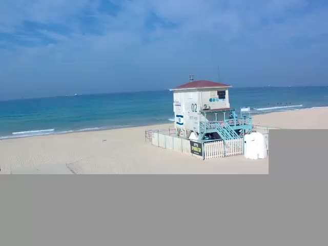 A multi-story white and blue building with a red roof, resembling a lifeguard station, stands on a wide sandy beach beside a calm turquoise ocean under a bright, partly cloudy sky.