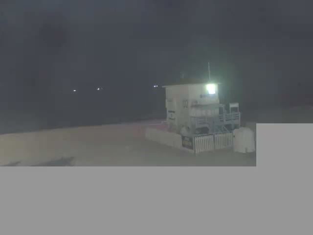 A multi-story white and blue building with a red roof, resembling a lifeguard station, stands on a wide sandy beach beside a calm turquoise ocean under a bright, partly cloudy sky.