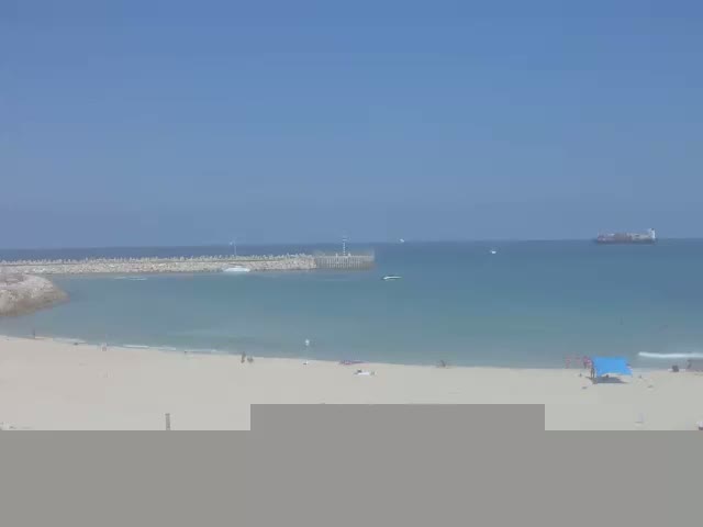 A sandy beach with a few people on it stretches out to a calm, light blue ocean with a breakwater and a distant cargo ship visible under a clear blue sky.