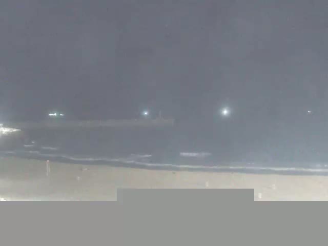 A dark, hazy night scene shows a beach with gentle waves lapping the shore, and a distant structure illuminated by faint lights.