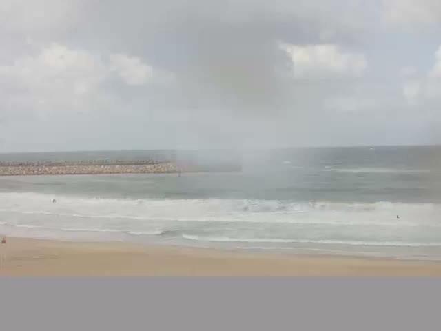 A sandy beach with white waves breaking on the shore leads to a hazy ocean featuring a distant rock breakwater, all beneath an overcast and cloudy sky.