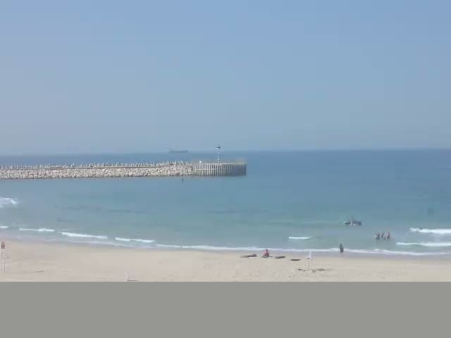 A sandy beach with white waves breaking on the shore leads to a hazy ocean featuring a distant rock breakwater, all beneath an overcast and cloudy sky.