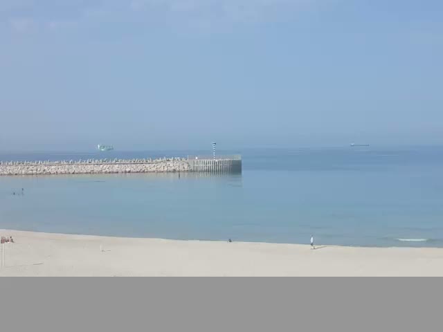 A tranquil beach scene on a clear, sunny day features a sandy shore meeting calm blue waters, a stone breakwater extending into the sea with a small beacon, and a few distant ships.