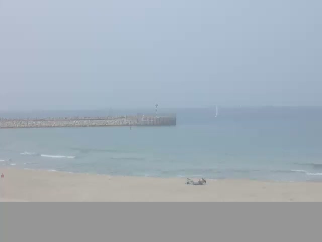 On a hazy and overcast day, a sandy beach with gentle waves leads to a body of water featuring a long stone breakwater with a lighthouse, a distant sailboat, and two people relaxing on the sand.