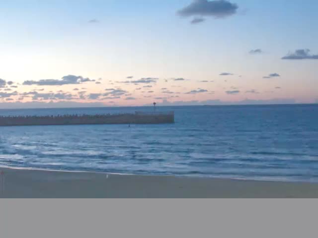 A serene view of a sandy beach and a breakwater extending into the calm blue ocean under a partly cloudy twilight sky.
