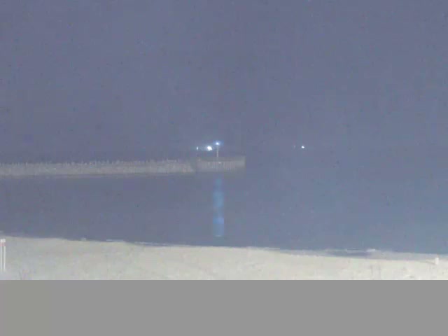 A dark night scene captures a sandy beach transitioning into calm water, with a distant pier or breakwater faintly lit under a clear or nearly clear sky.