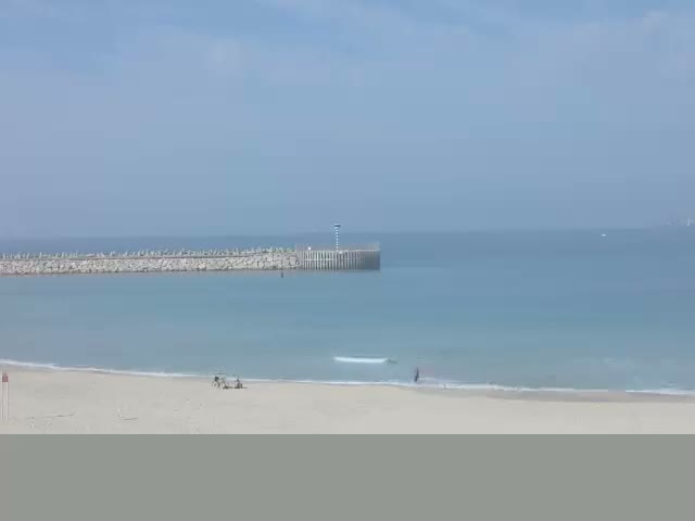 A sunny day at a sandy beach features calm blue water with a long rock breakwater extending into the sea, where a few people are visible enjoying the shore.