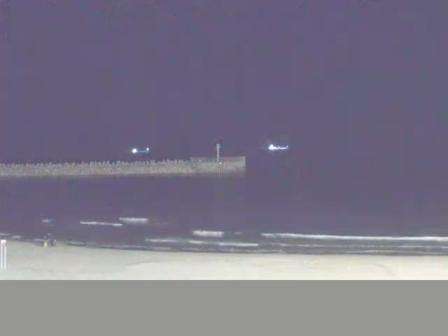 A sunny day at a sandy beach features calm blue water with a long rock breakwater extending into the sea, where a few people are visible enjoying the shore.