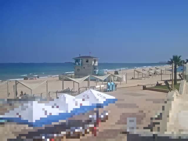 A sandy beach with calm waves under a clear blue sky features several shaded beach areas, a lifeguard station, and a few palm trees.