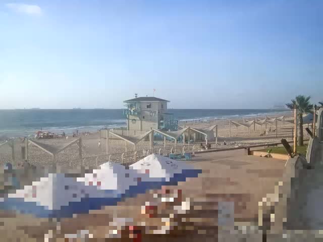 A sandy beach with calm waves under a clear blue sky features several shaded beach areas, a lifeguard station, and a few palm trees.