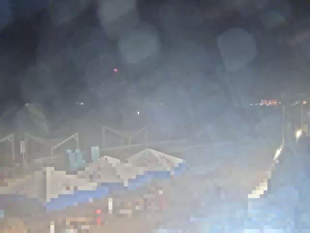 A hazy night scene shows a collection of light-colored tents or structures, possibly at an outdoor event, with indistinct figures and lights in the background.