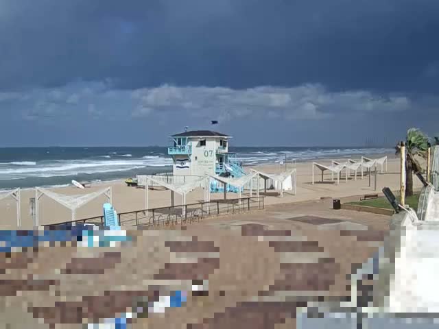 A beach with a blue and white lifeguard tower, numerous white-canopied structures, and an ocean with white-capped waves is visible under a dramatic, heavily overcast sky filled with dark clouds.