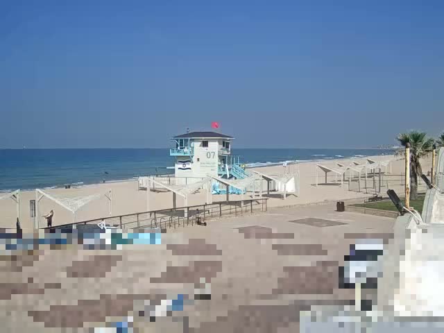 A beach with a blue and white lifeguard tower, numerous white-canopied structures, and an ocean with white-capped waves is visible under a dramatic, heavily overcast sky filled with dark clouds.