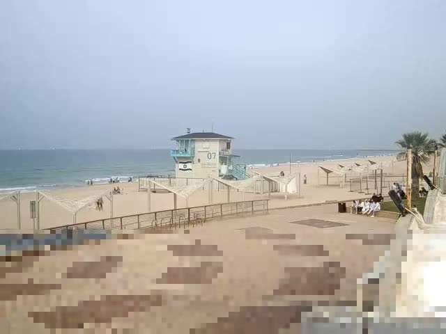 An overcast day at a sandy beach features a prominent white and blue lifeguard tower overlooking the ocean, with several people walking and sitting on the sand and a paved boardwalk, and palm trees visible on the right.