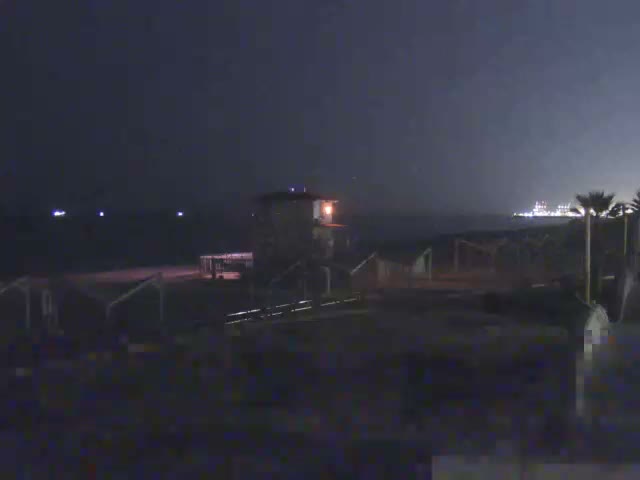 A sunny day at a sandy beach features a white and blue lifeguard tower, multiple open-air shelters, a calm blue sea with distant boats, and a paved promenade in the foreground.