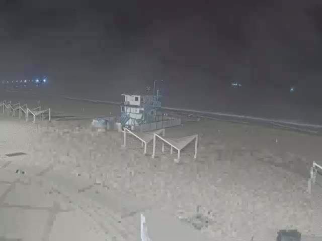 A nighttime beach scene shows a small building and several beachside structures under a hazy sky.