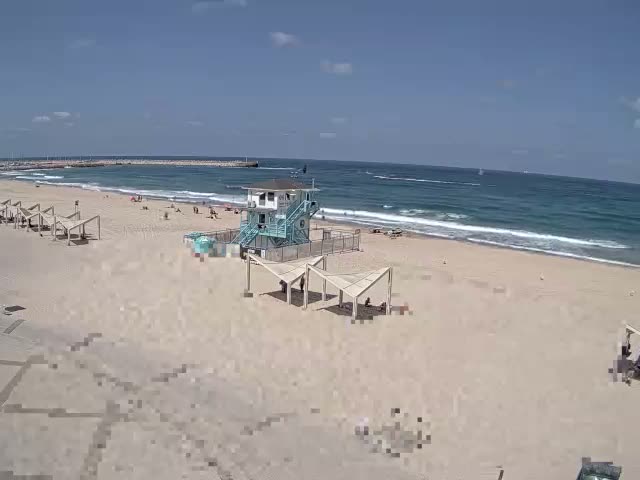 A sandy beach with a lifeguard stand and several shaded structures is shown on a sunny day with light waves rolling onto the shore.