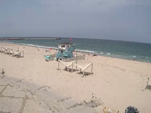 A sandy beach with a lifeguard station and several shaded structures is shown on a partly sunny day with light waves lapping the shore.