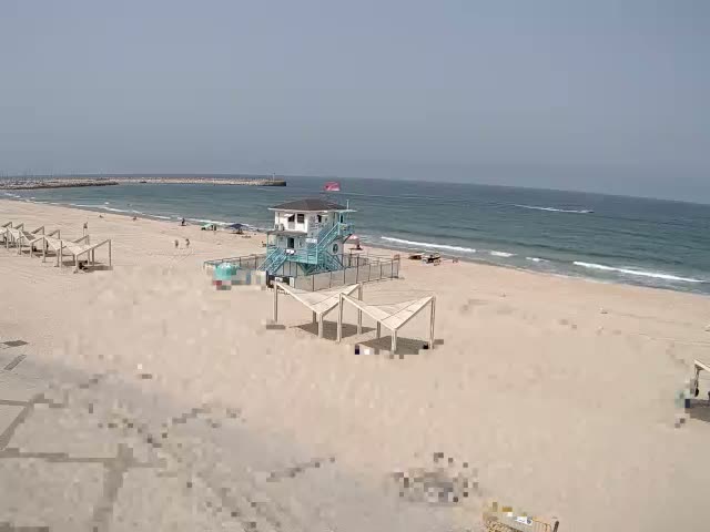 A sandy beach with a lifeguard station, several shaded structures, and a few people is visible on a sunny day, with a calm sea in the background.