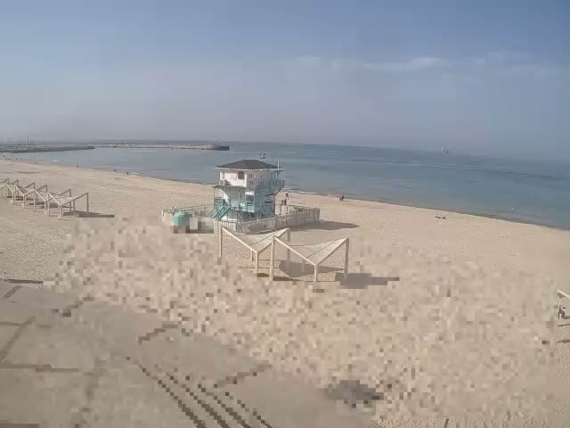 A wide sandy beach featuring a two-story lifeguard station and several shaded structures is visible on a clear, sunny day with calm blue water, a distant harbor, and a few people enjoying the outdoor conditions.