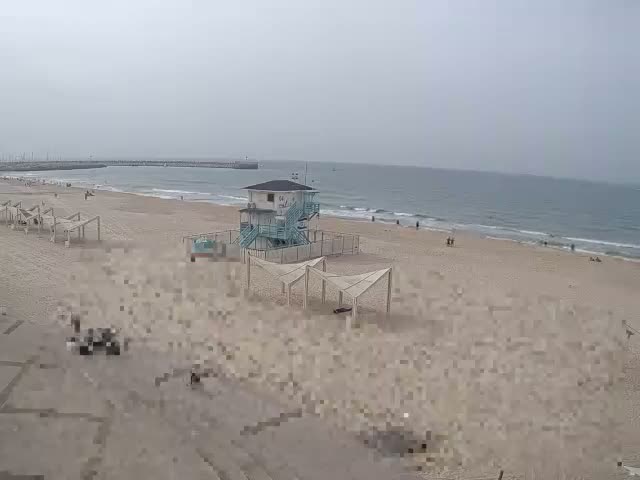 On an overcast day, a sandy beach features a light blue lifeguard tower, multiple open-air shade structures, a long pier extending into the sea, and scattered people walking near the water's edge.