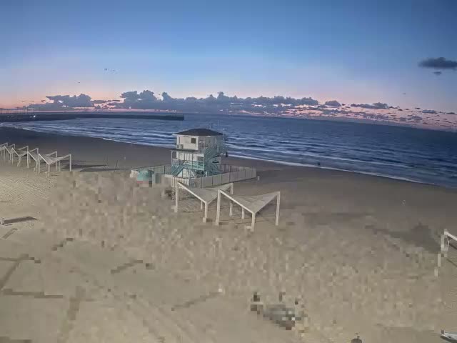 A sandy beach with a turquoise-trimmed lifeguard tower and several white shade structures leads to calm ocean waters under a twilight sky with scattered clouds showing hues of deep blue, purple, and orange, with a distant pier visible on the left.