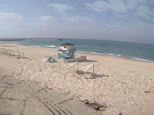 A wide sandy beach stretches towards a blue ocean with gentle waves under a bright, partially cloudy sky, featuring a prominent white and blue lifeguard tower and several wooden shade structures on the sand, with a distant breakwater.