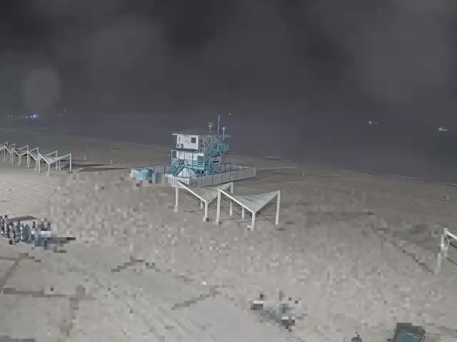 A wide sandy beach stretches towards a blue ocean with gentle waves under a bright, partially cloudy sky, featuring a prominent white and blue lifeguard tower and several wooden shade structures on the sand, with a distant breakwater.