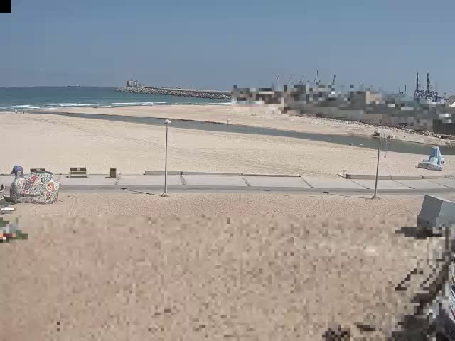 A sandy beach stretches towards a calm ocean under a clear, sunny sky, with a harbor visible in the distance.