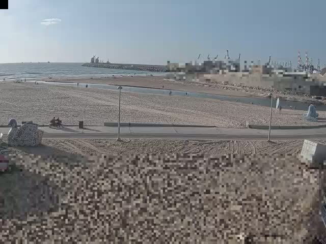 A sandy beach stretches towards a calm ocean under a clear, sunny sky, with a harbor visible in the distance.