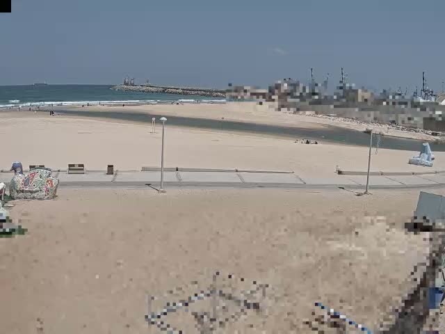A sandy beach with a calm ocean under sunny skies shows a few people relaxing near a waterway that leads to a harbor.