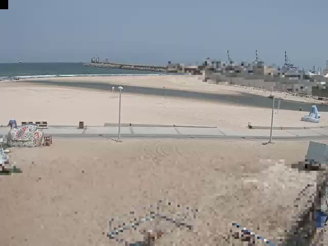 A sandy beach with a calm ocean and a harbor in the background on a sunny day.