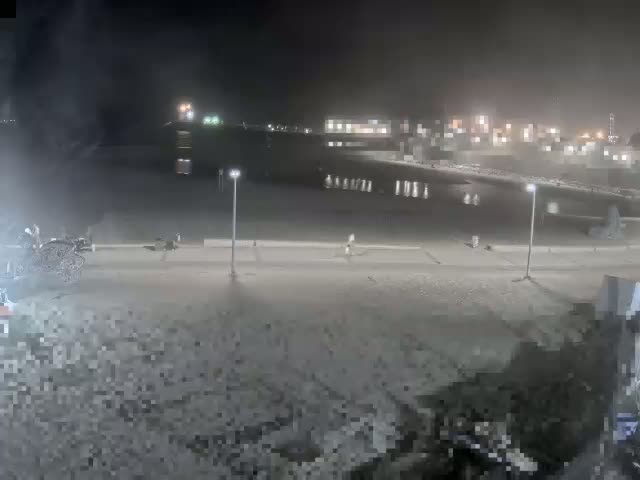 A nighttime view shows a mostly empty paved area near a body of water under hazy or foggy conditions, with a few people and a horse-drawn carriage visible.