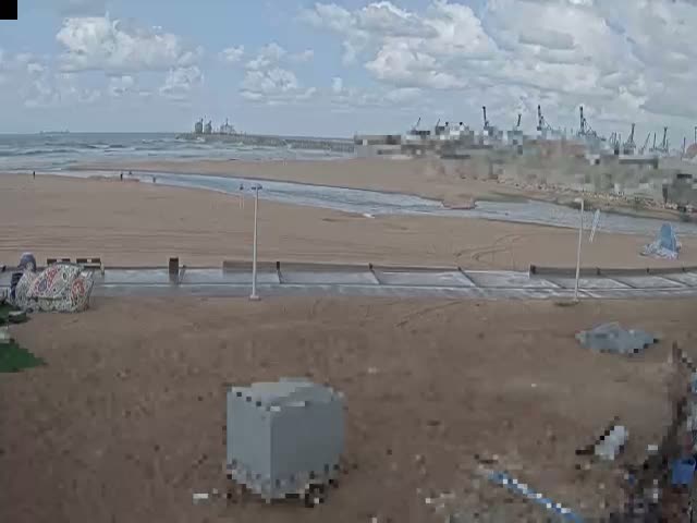 A partly cloudy sky hangs over a wide sandy beach where a waterway meets the ocean, bordered by a paved path and featuring distant industrial port cranes and a few scattered people.