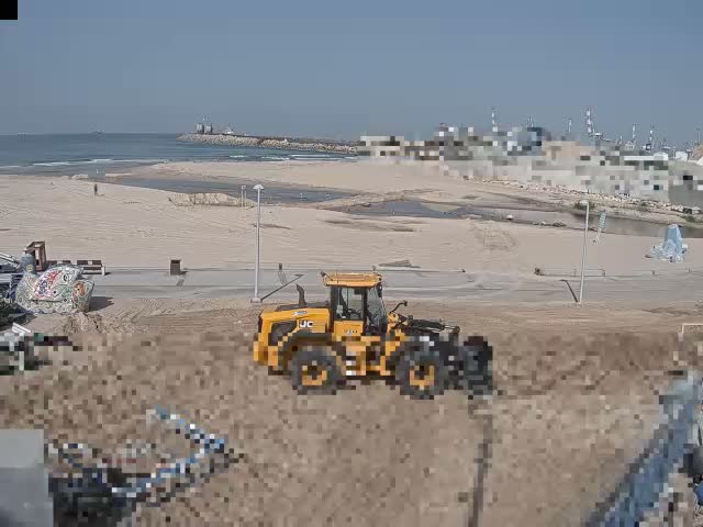 A partly cloudy sky hangs over a wide sandy beach where a waterway meets the ocean, bordered by a paved path and featuring distant industrial port cranes and a few scattered people.