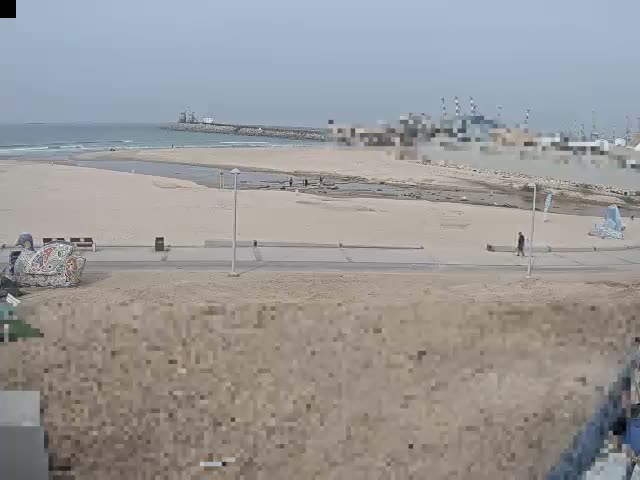 On an overcast day, a wide sandy beach with a stream flowing into the calm sea is visible, bordered by a paved path with a few pedestrians, and a long breakwater with distant industrial structures extends into the water.