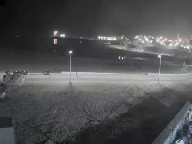 A blurry, low-light image captures a rough, possibly snow-dusted or sandy ground next to a road with two streetlights, leading towards a distant cityscape or industrial complex whose numerous blurred lights reflect on a dark horizon under a hazy night sky.