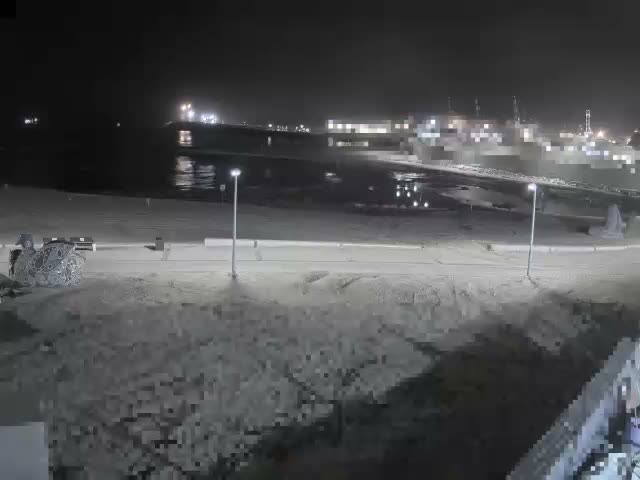 On a fair, partly cloudy day, a coastal view features a wide sandy beach where a river meets the sea, with a distant pier, two electric scooters on a paved path, and a colorful mosaic sculpture in the foreground, all framed by distant industrial structures.