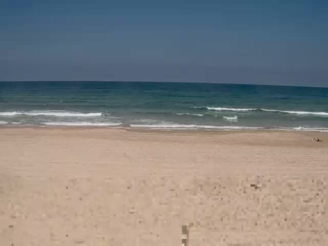A wide sandy beach with small waves rolling onto the shore under a clear blue sky.
