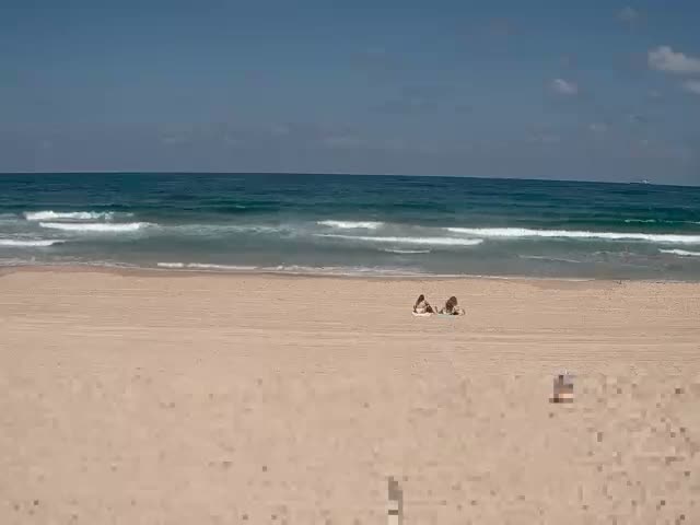 Two people lie on a sandy beach under a mostly sunny sky, with gentle waves rolling in on a calm ocean.
