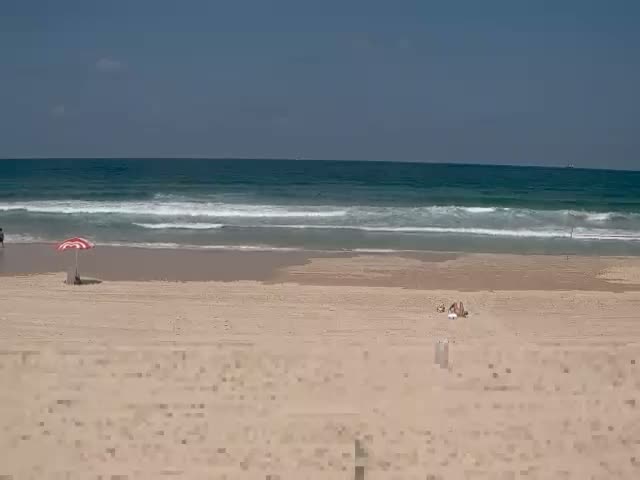 A sandy beach with a few people sunbathing and a single red and white striped umbrella is visible under a clear sky with a calm sea showing small waves.