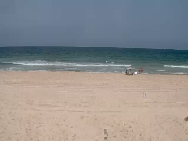 A sandy beach with a few people under an umbrella near the gently lapping ocean waves on a clear day.
