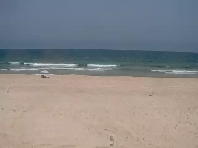 A mostly empty sandy beach with small waves under a clear sky shows a single beach umbrella and two lounge chairs.
