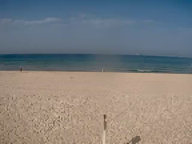 A wide sandy beach under a clear, sunny sky with scattered clouds meets a calm blue ocean featuring distant ships, as a few people walk near the shoreline.